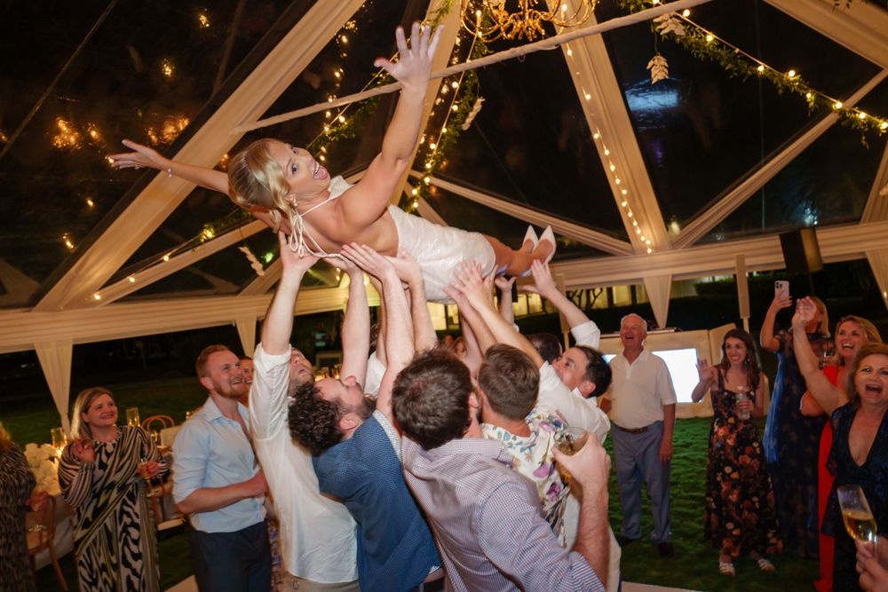 Wedding celebration with guests lifting someone in the air under a decorated tent ceiling.