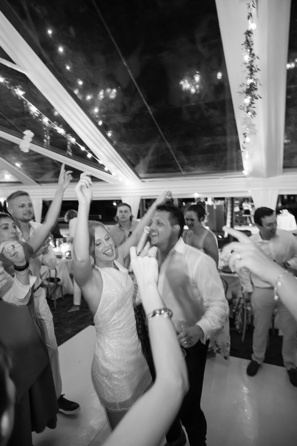 Wedding party celebrates with dancing and raised arms under a clear-roofed tent decorated with lights.