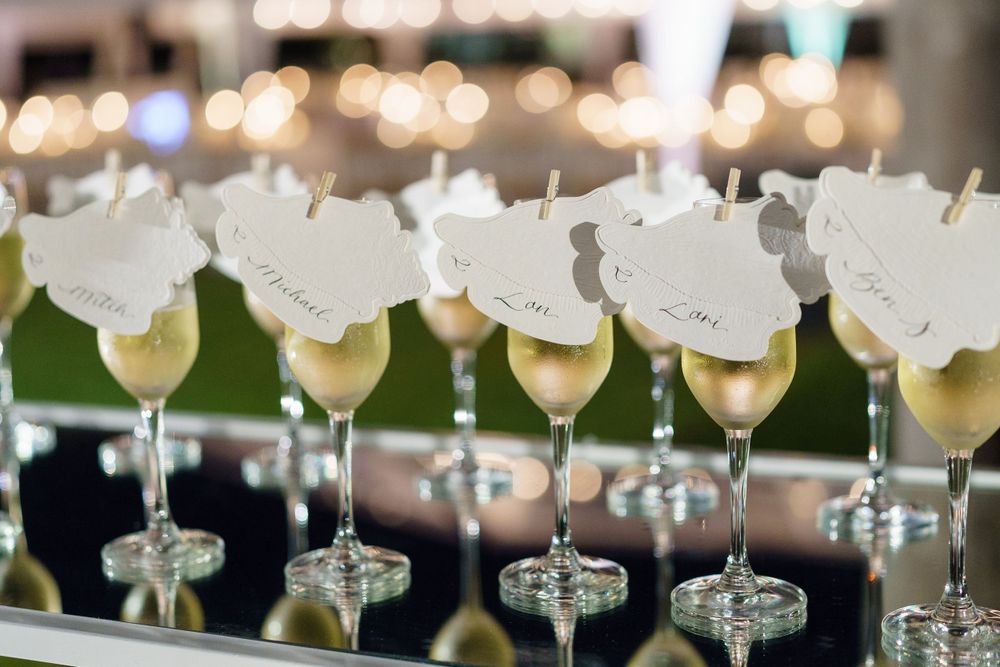 Row of elegant cocktail glasses with white floral garnishes lined up on a mirrored surface with bokeh lights in background.