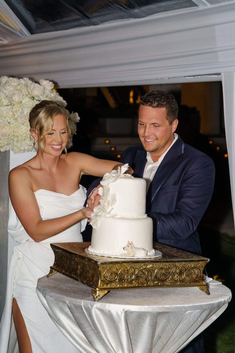 Newlyweds cut their elegant white wedding cake displayed on an ornate gold stand at their reception.