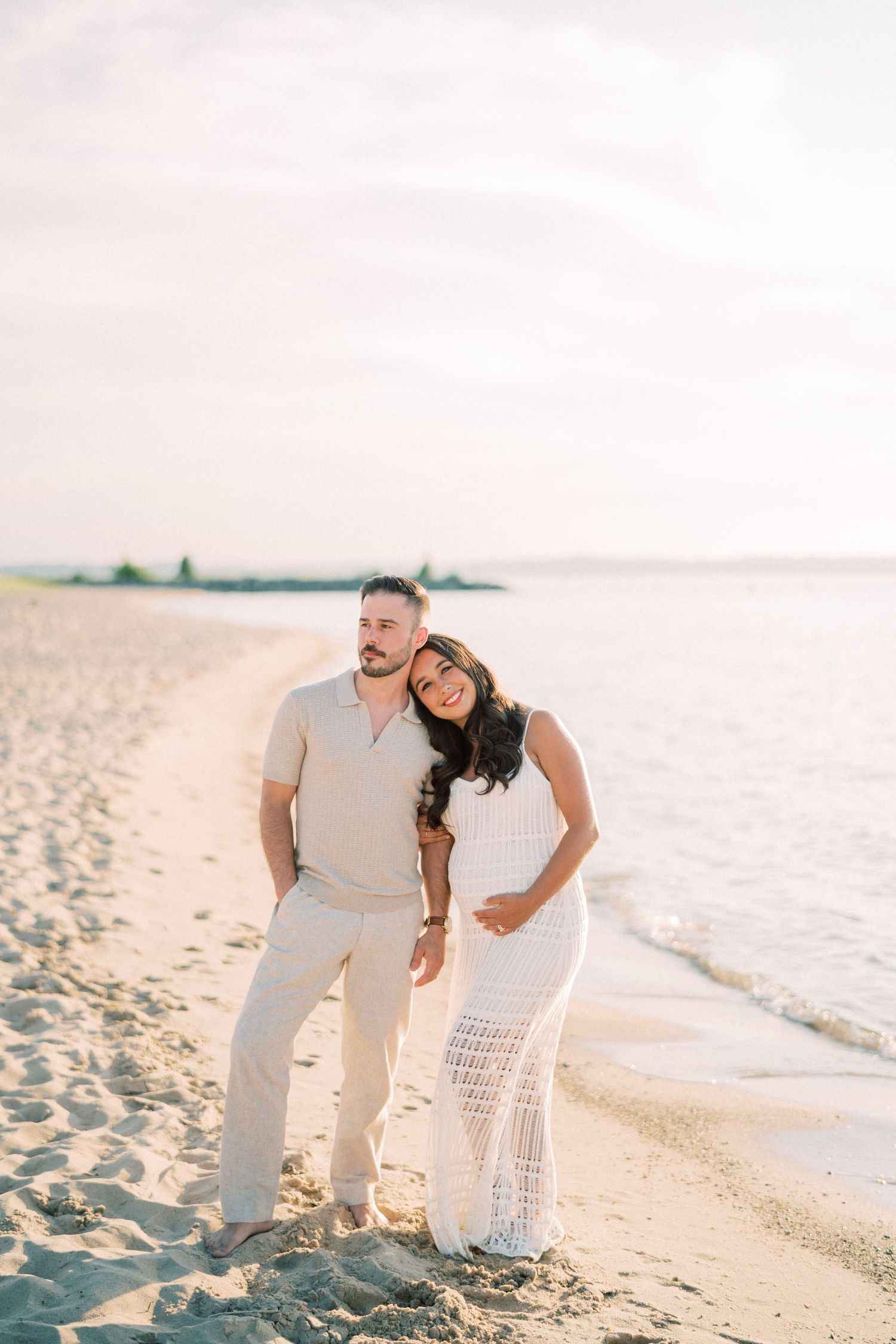 A couple in white outfits poses together on a sandy beach at sunset.