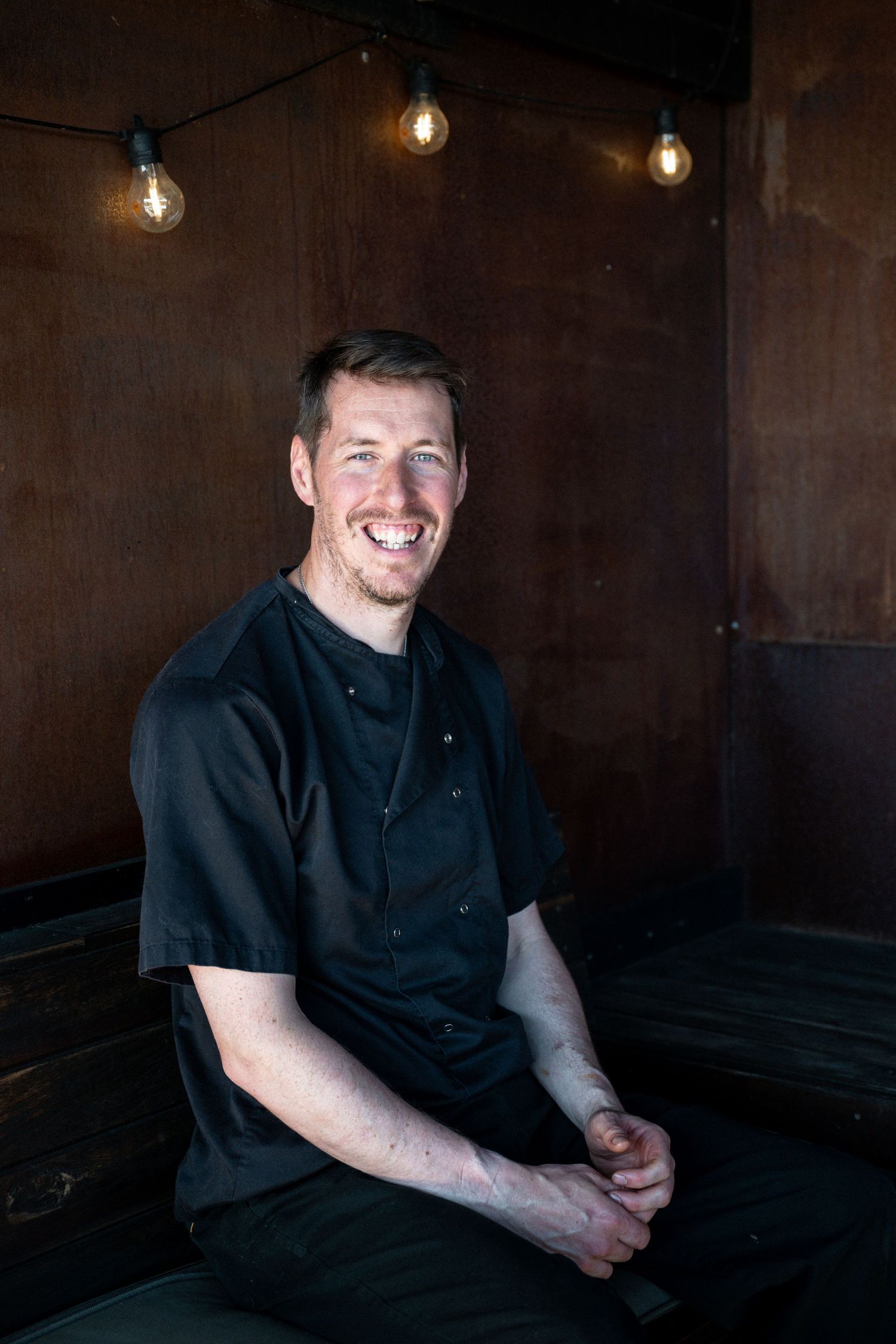 Chef in black uniform smiling while sitting in dimly lit restaurant with hanging pendant lights.