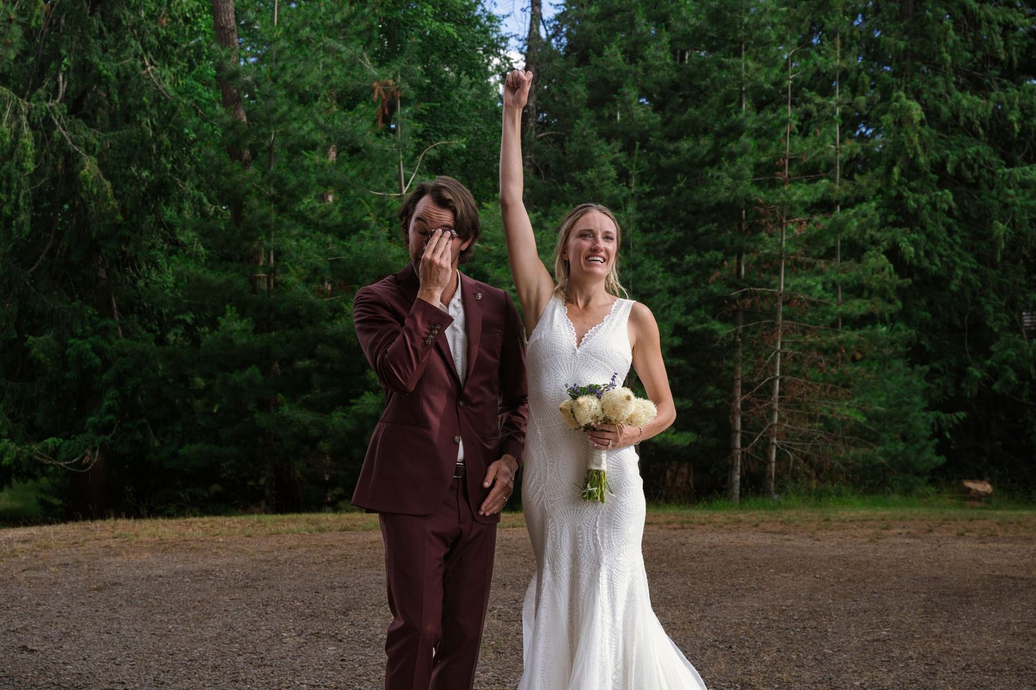 A wedding couple shares a romantic moment surrounded by tall evergreen trees in a rustic outdoor setting.