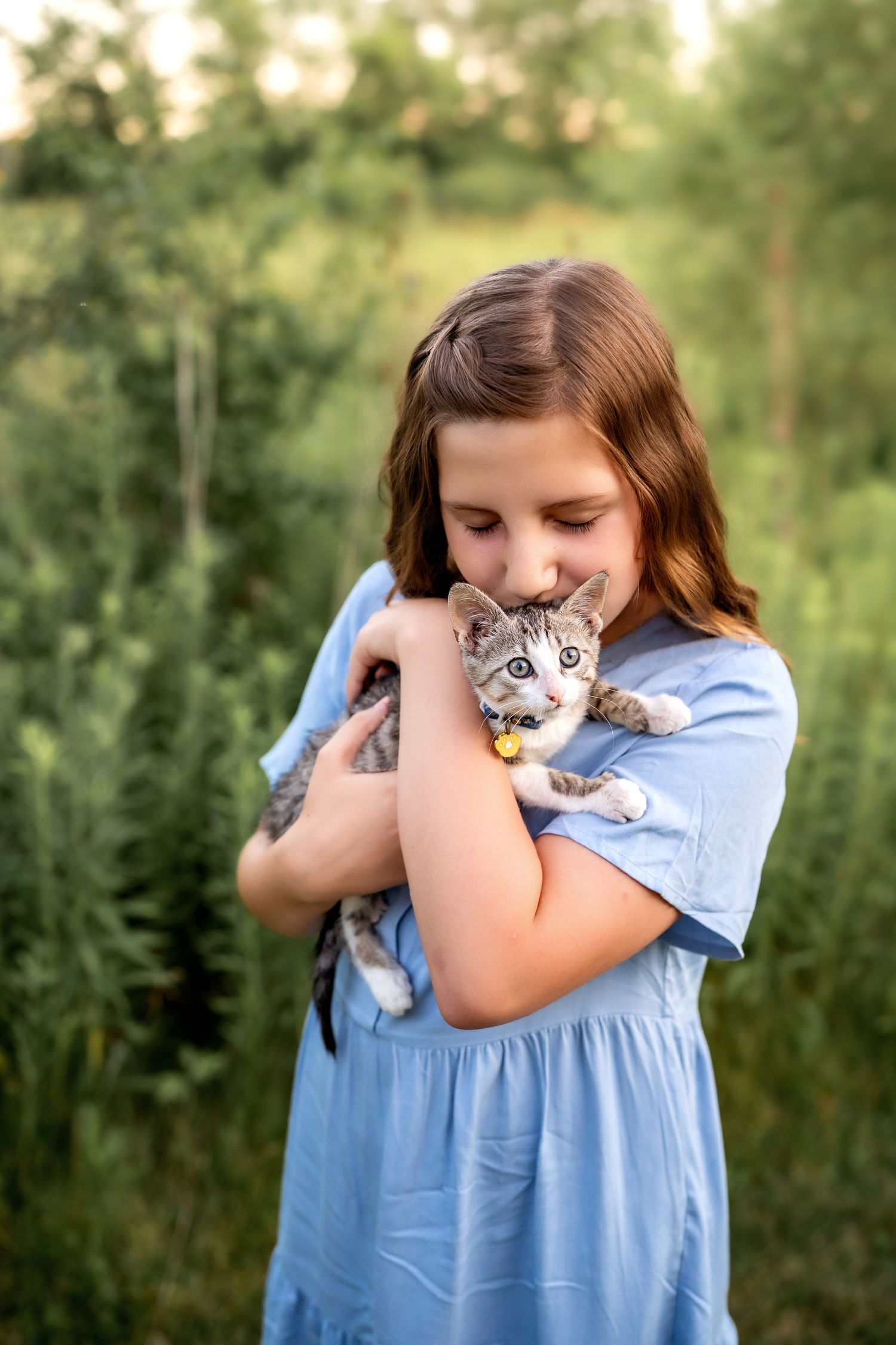 Someone wearing a light blue dress snuggles with a small gray tabby cat against a green outdoor background.