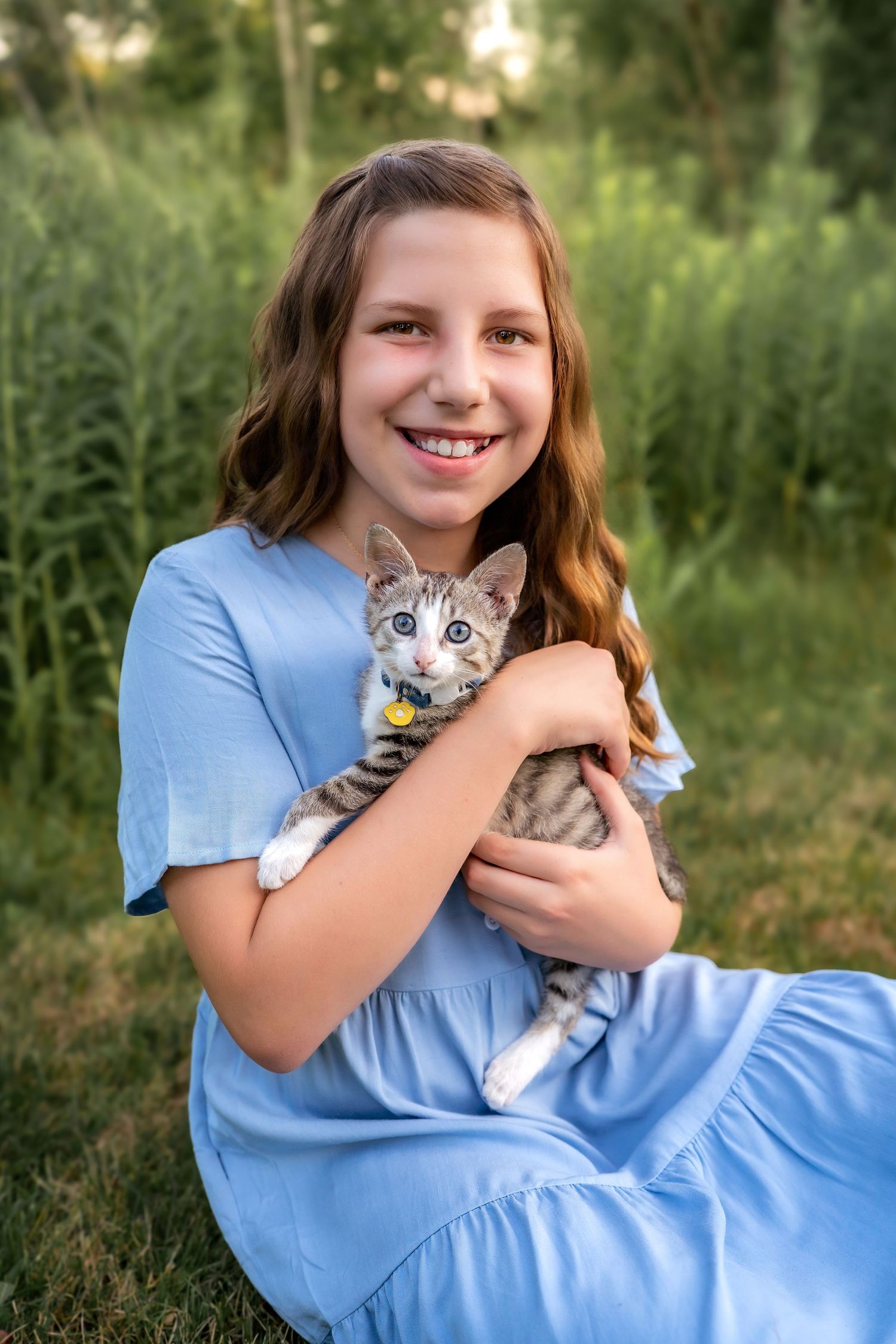 A young person in a blue dress sits in a grassy field while holding a gray tabby kitten in their arms.