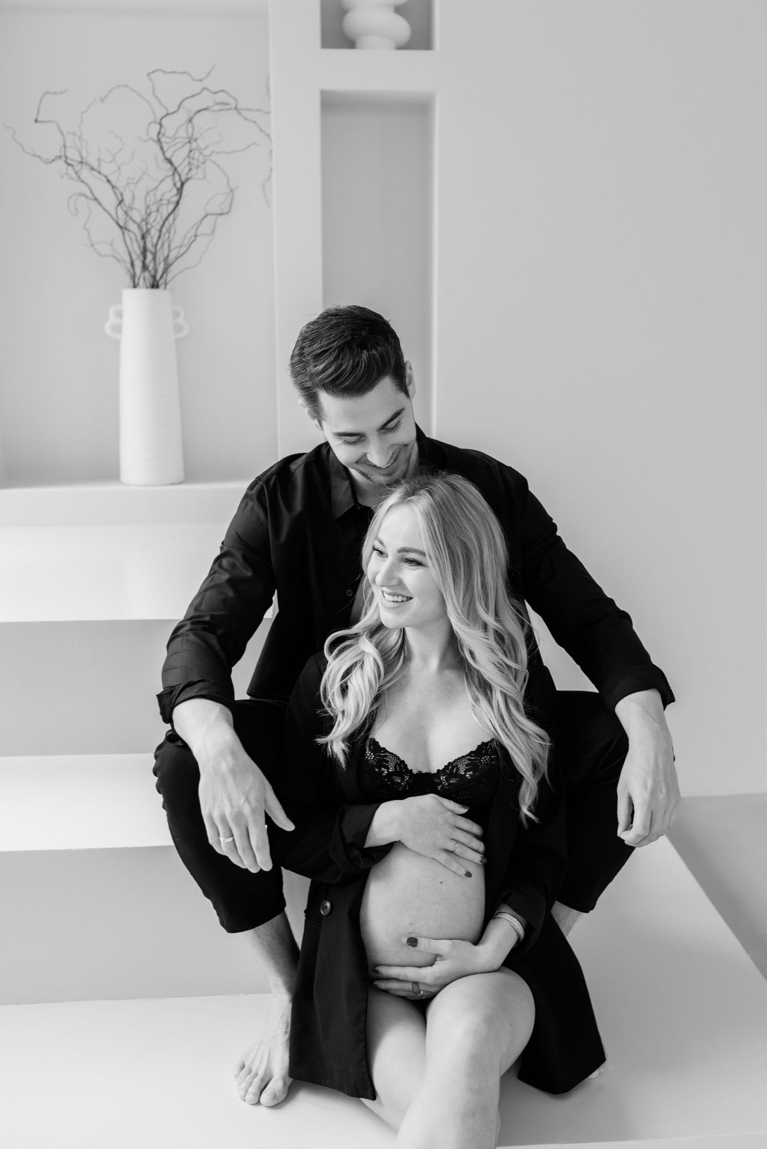 Black and white maternity photo showing intimate couple sitting together with bare pregnant belly in modern setting.