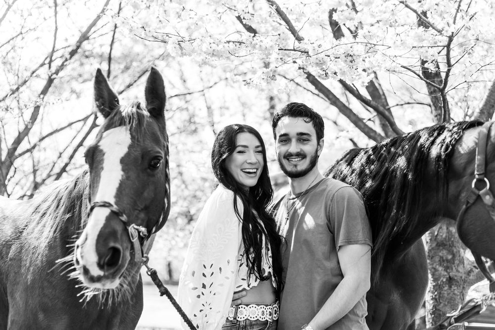 Black and white photo of a couple smiling with horses amid blooming trees.