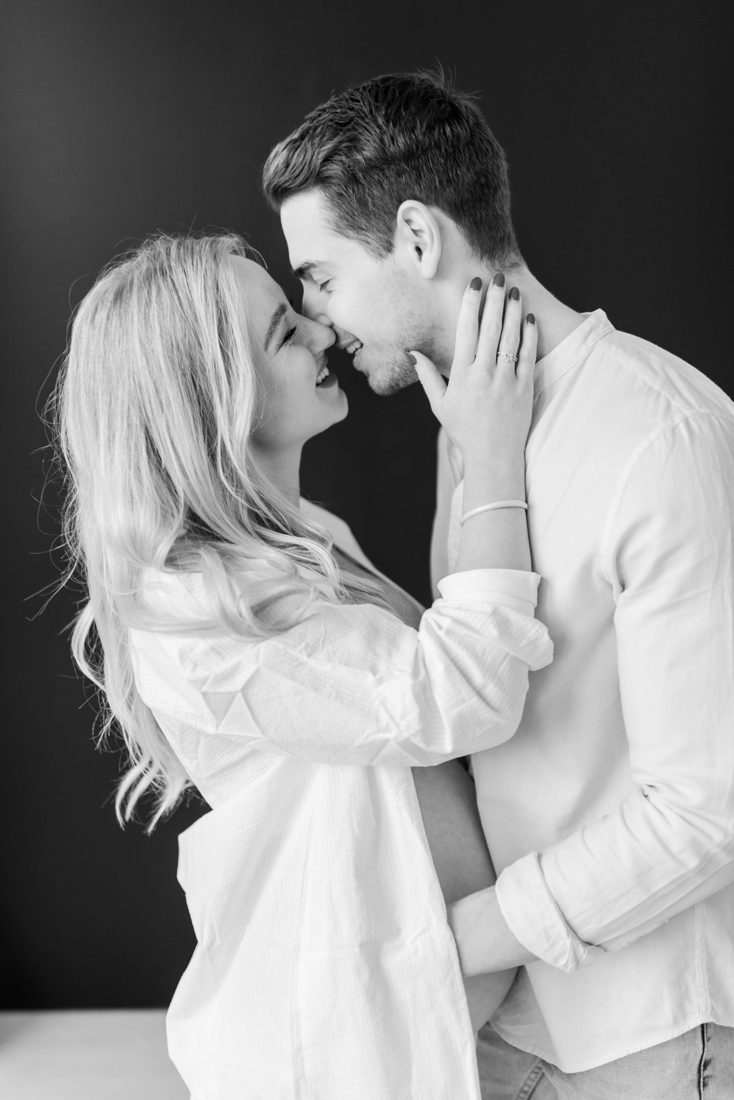 Intimate black and white portrait of a couple in white clothing embracing and touching foreheads against dark background.