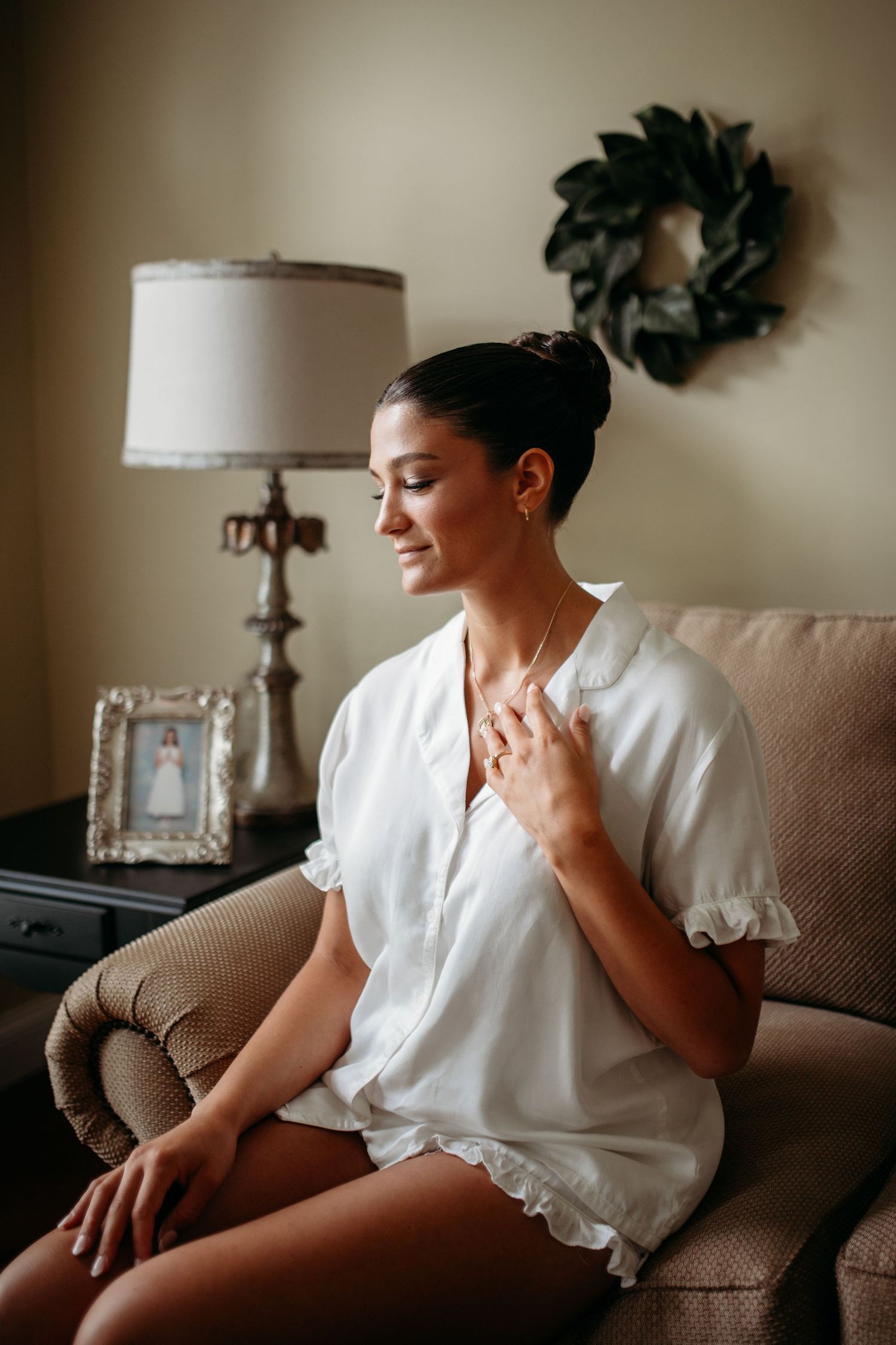 A Catholic bride sitting sitting reflectively on a brown couch in warm natural lighting, creating a serene mood prior to her wedding day.