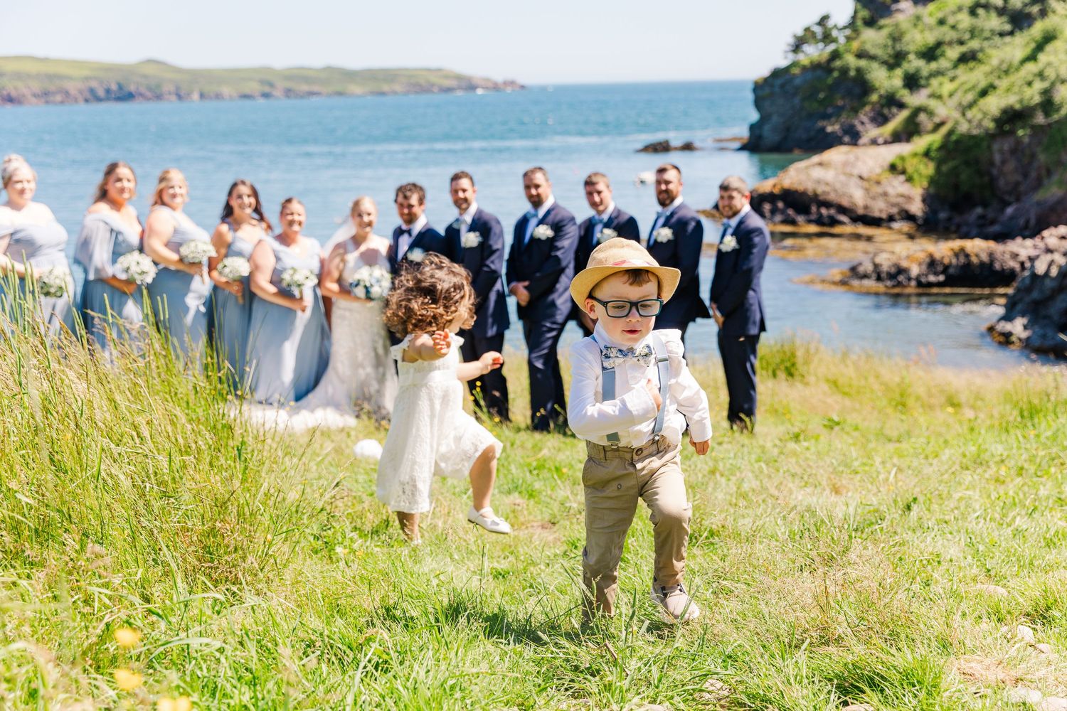 Bridal party gathers on a scenic coastal hillside with grassy slopes and blue ocean waters in the background.