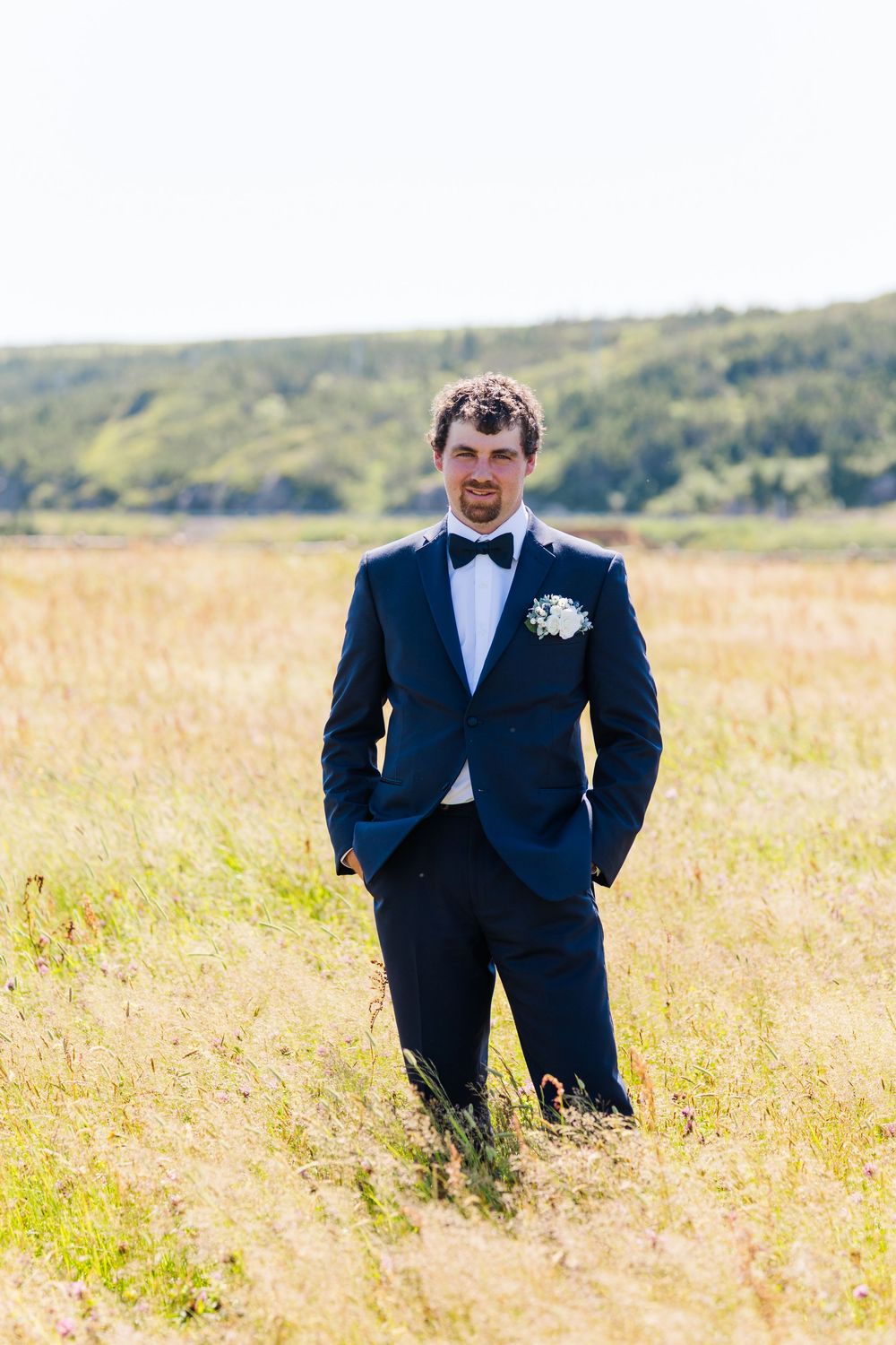 A groom in a navy blue suit and bow tie stands in a sunlit field of tall grass with hills in the background.