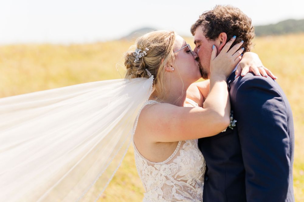 A joyful newlywed couple embraces in a sun-drenched field with the bride's veil flowing in the breeze.