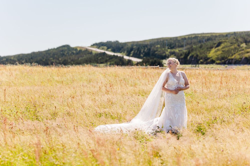 Two people in wedding attire walk through a sun-drenched golden grass field with rolling hills in the background.