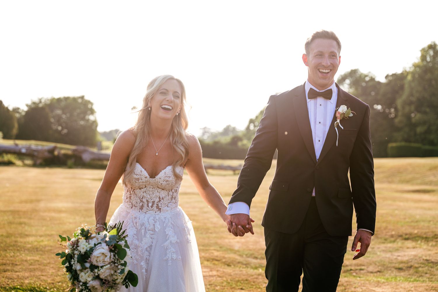 Newlywed couple walking and holding hands in golden sunlight on grassy field with bouquet.