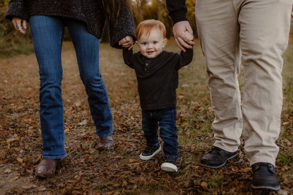 A toddler in a black sweater holds hands with adults while walking on fallen autumn leaves.