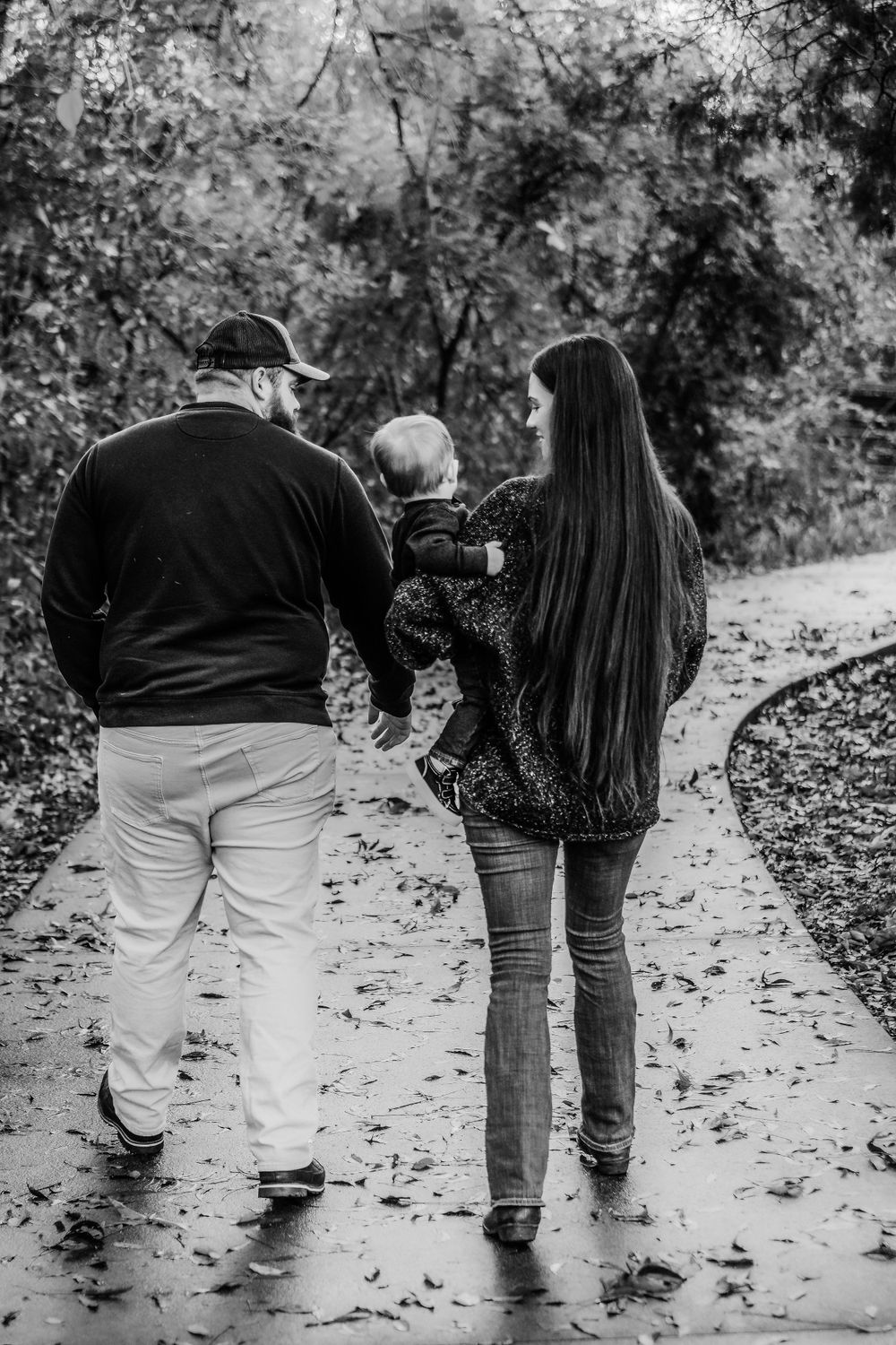 A couple walks along a leaf-covered path holding a small child between them on an autumn day in black and white.