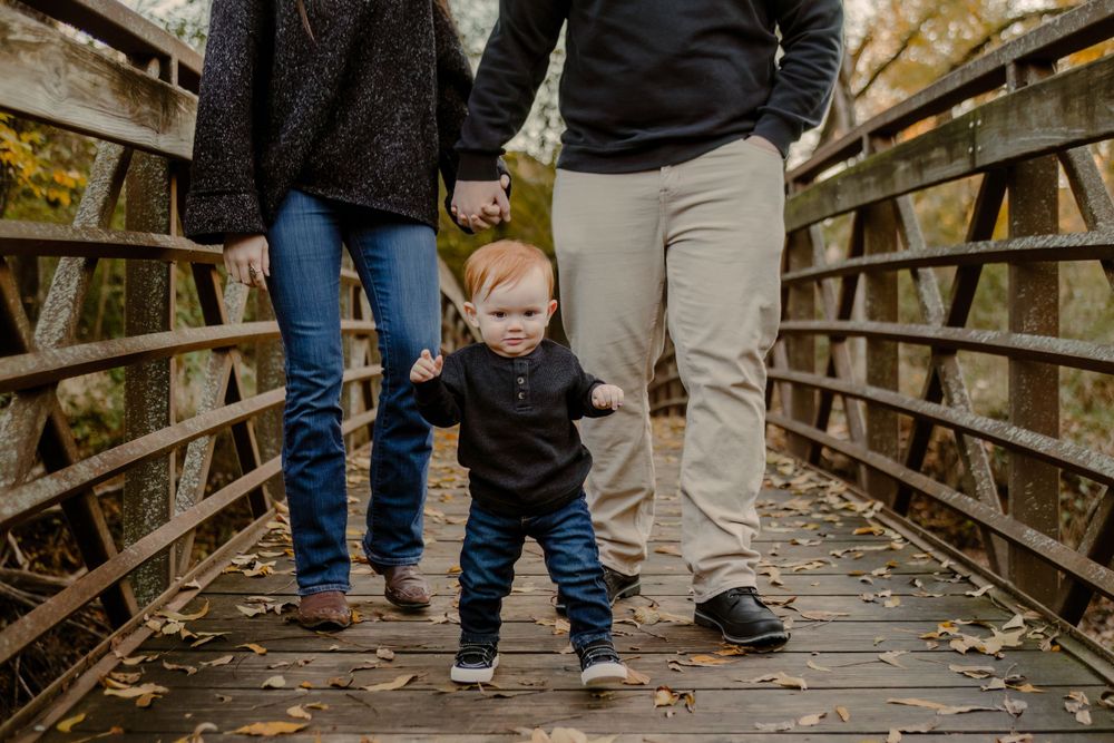 A small child in a black sweater stands between two adults on a wooden footbridge during autumn.