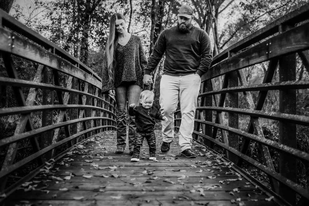 A family walks together holding hands across a wooden bridge covered in fallen autumn leaves in black and white.