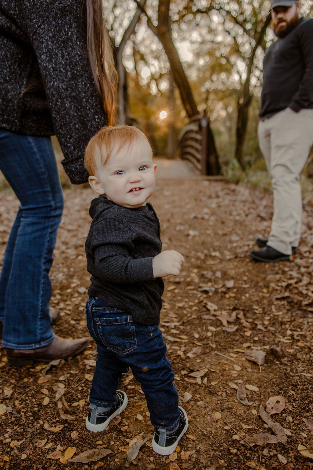A small child in jeans and a dark sweater smiles while walking on a leaf-covered trail during golden hour.