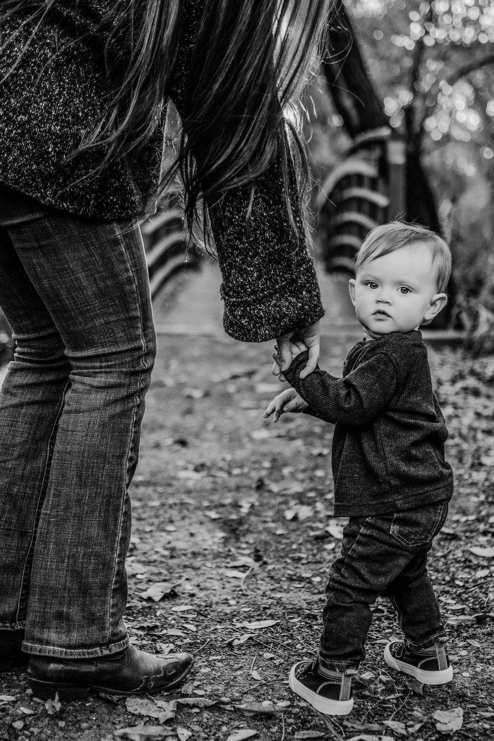 Black and white photo of a small toddler in dark clothing holding hands with an adult on a leaf-covered forest path.