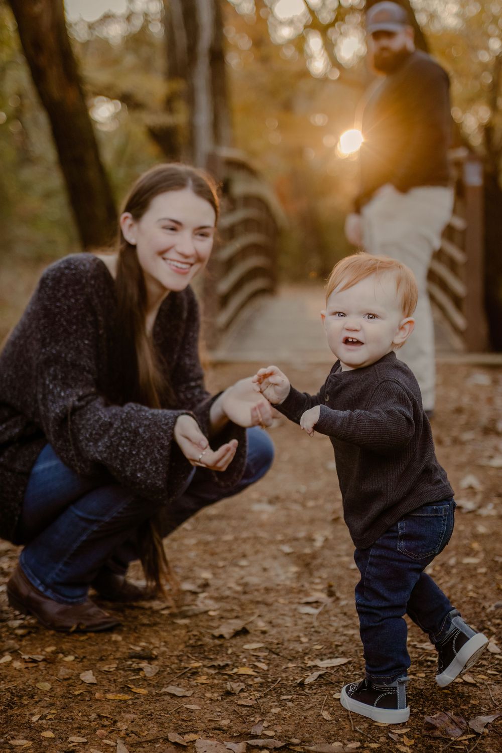 A toddler in jeans and sneakers plays on a wooded path at sunset while an adult crouches nearby.