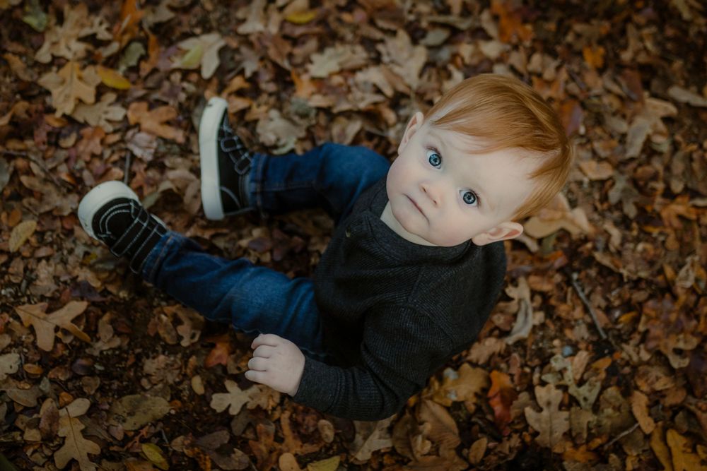 A child sits on autumn leaves wearing a dark sweater and blue jeans while looking up at the camera.