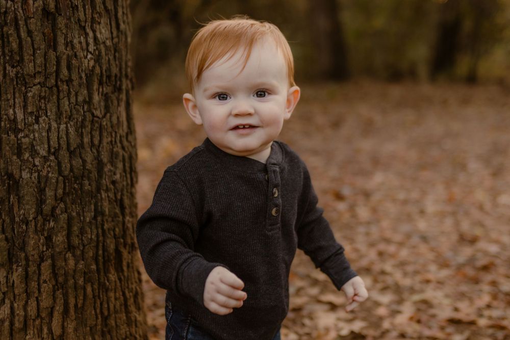 Small child in dark sweater smiles while standing next to tree trunk surrounded by autumn leaves.