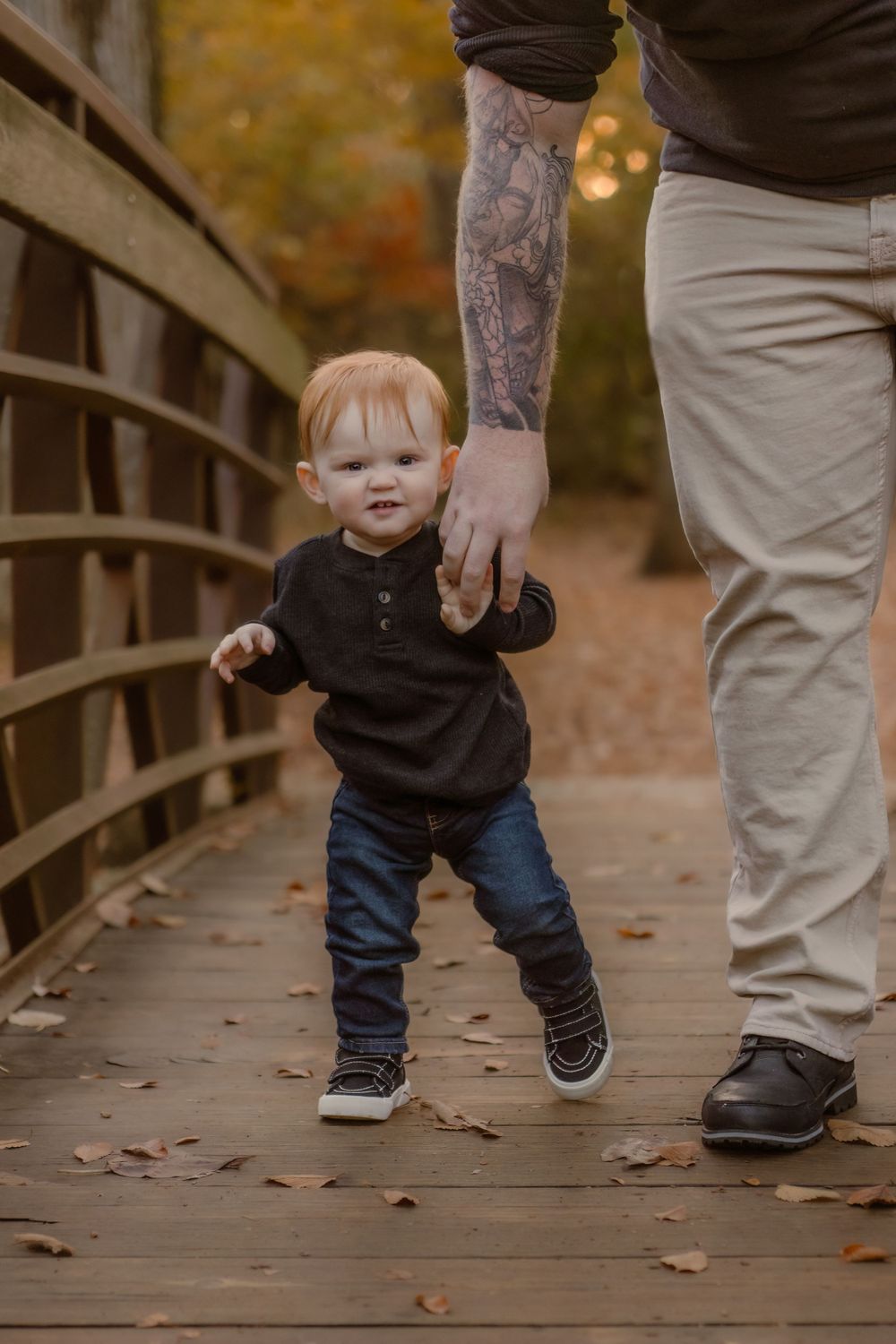 Small child holding hands with adult wearing tattoo sleeve while walking on wooden bridge during autumn.