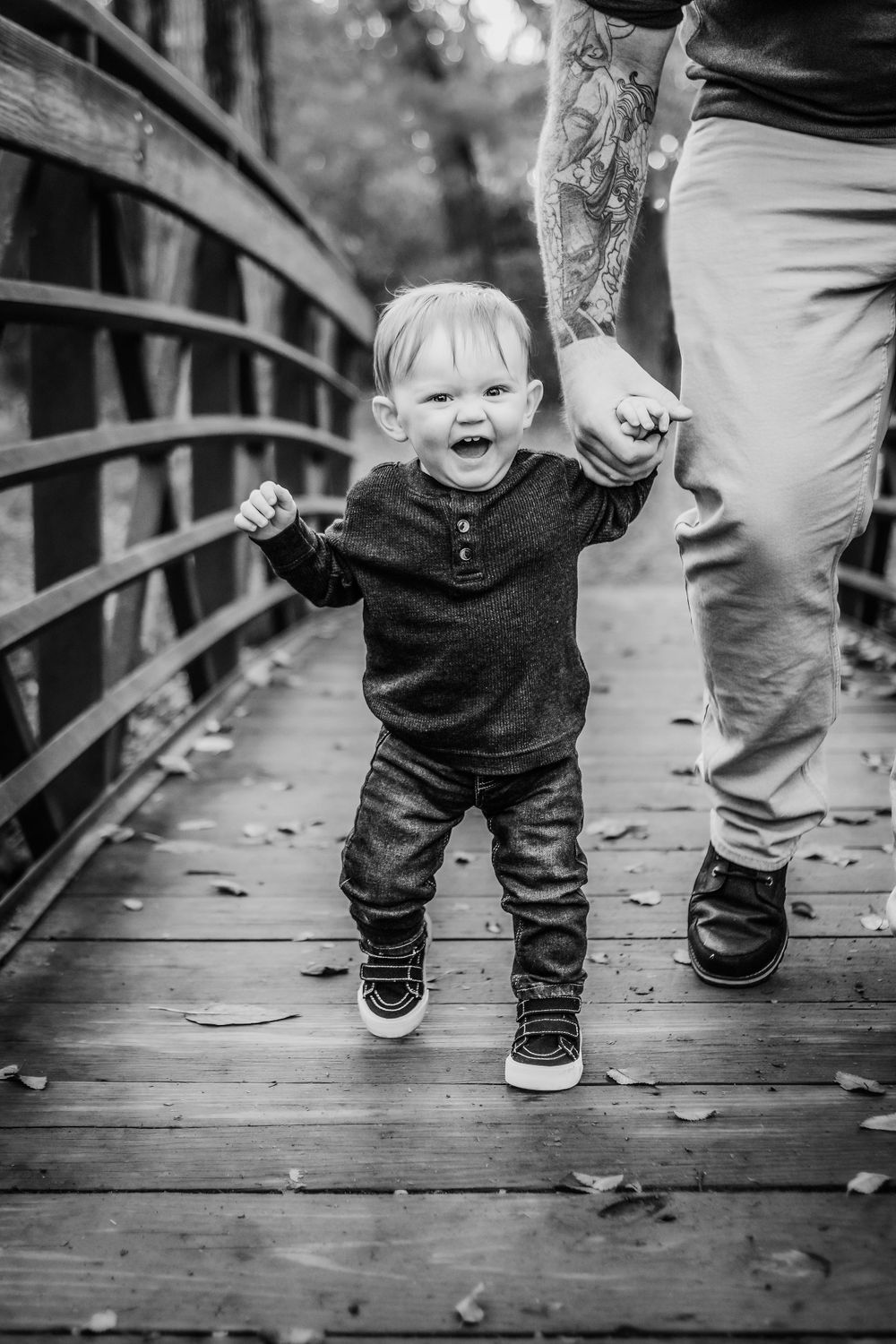 A joyful toddler walks on a wooden bridge while holding an adult's tattooed hand in black and white.