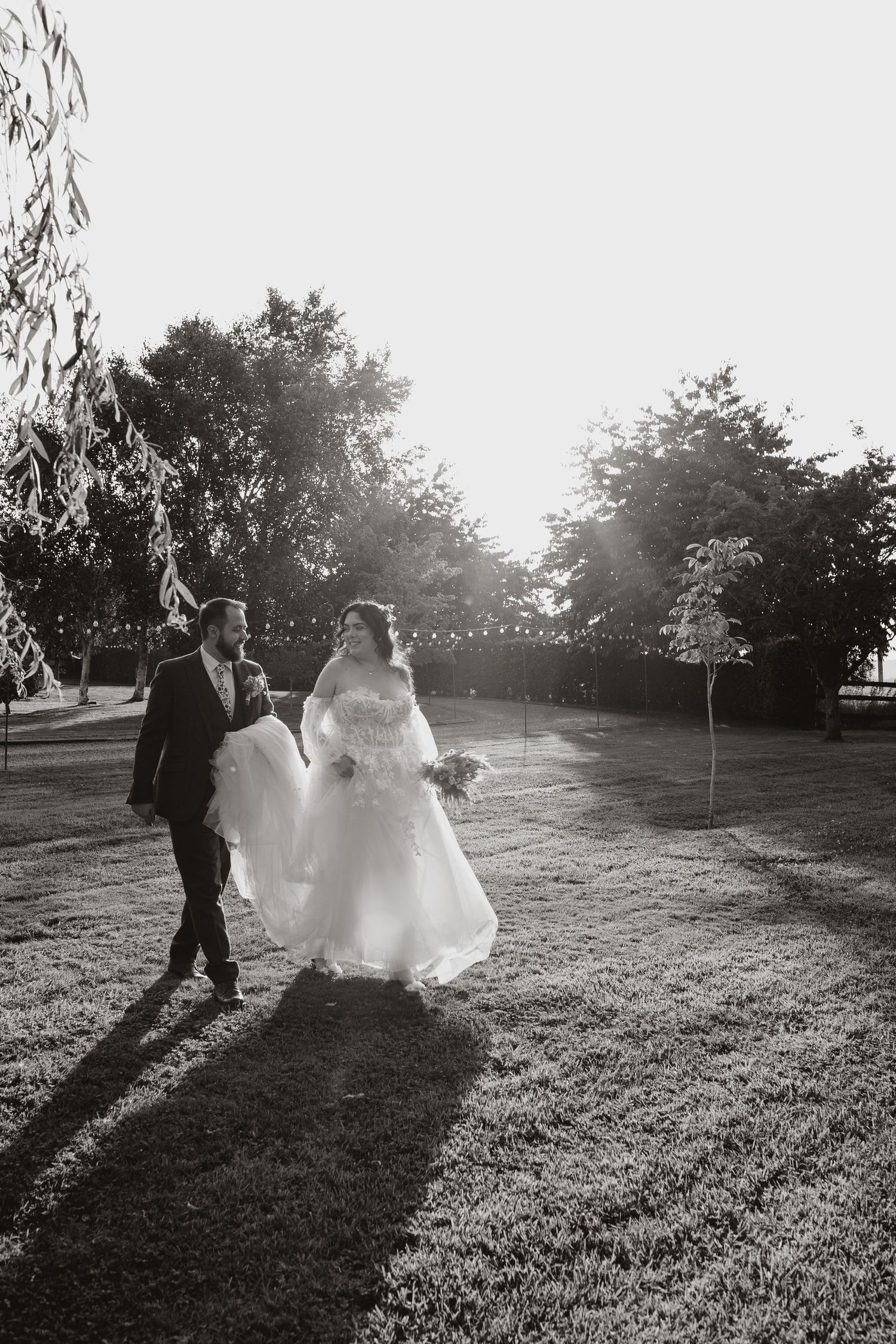 A couple walks on a sunlit lawn during their wedding day with string lights hanging overhead at sunset.