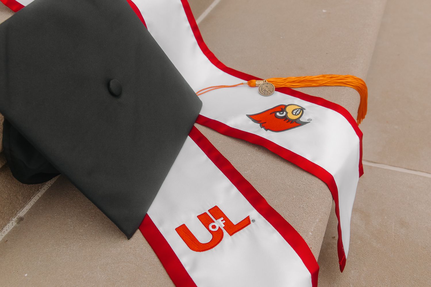 A black graduation cap and white stole with red trim and embroidered letters rests on a surface.