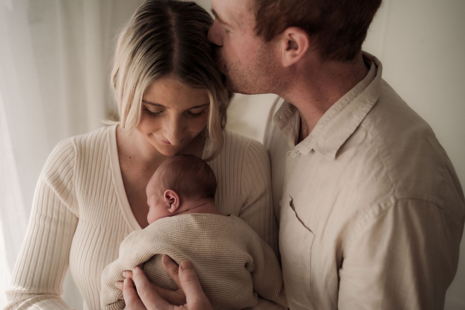 Newborn baby photo session with parents in neutral tones showing tender moments and cuddles in soft lighting.