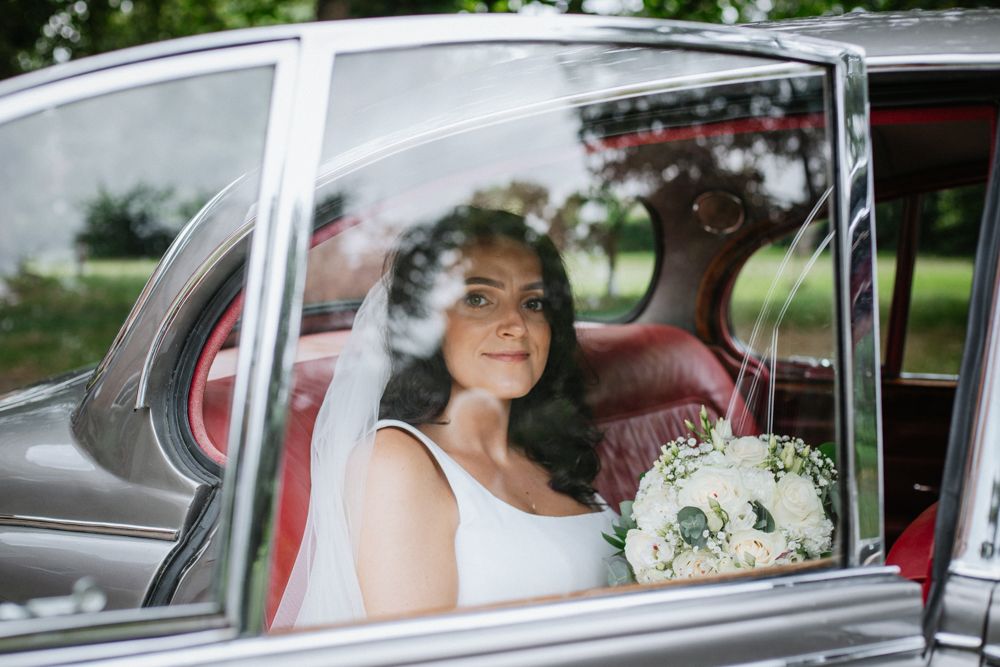A bride with white wedding dress and bouquet sitting inside a classic car with reflections on the window.
