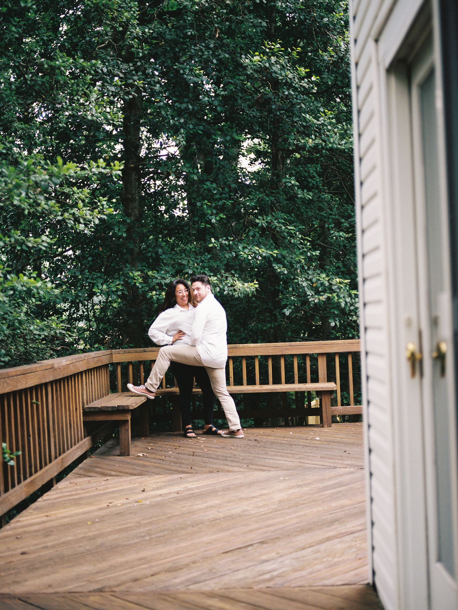 Romantic couple sharing tender moments on a wooden deck surrounded by lush green forest scenery.