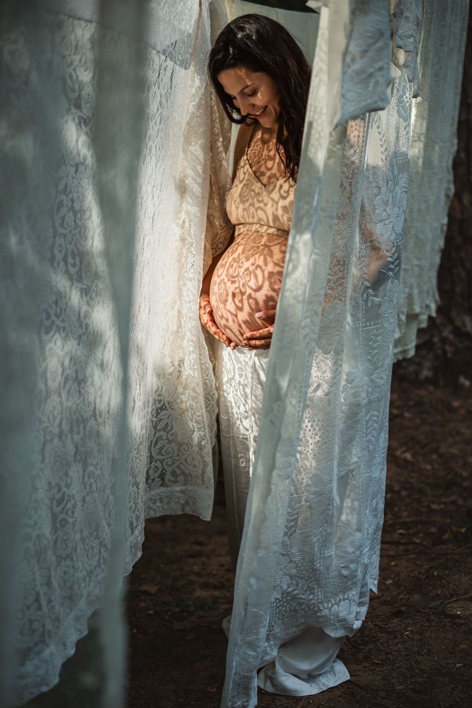 Beautiful maternity photo with sunlit lace curtain patterns casting shadows on pregnant belly in soft natural light.