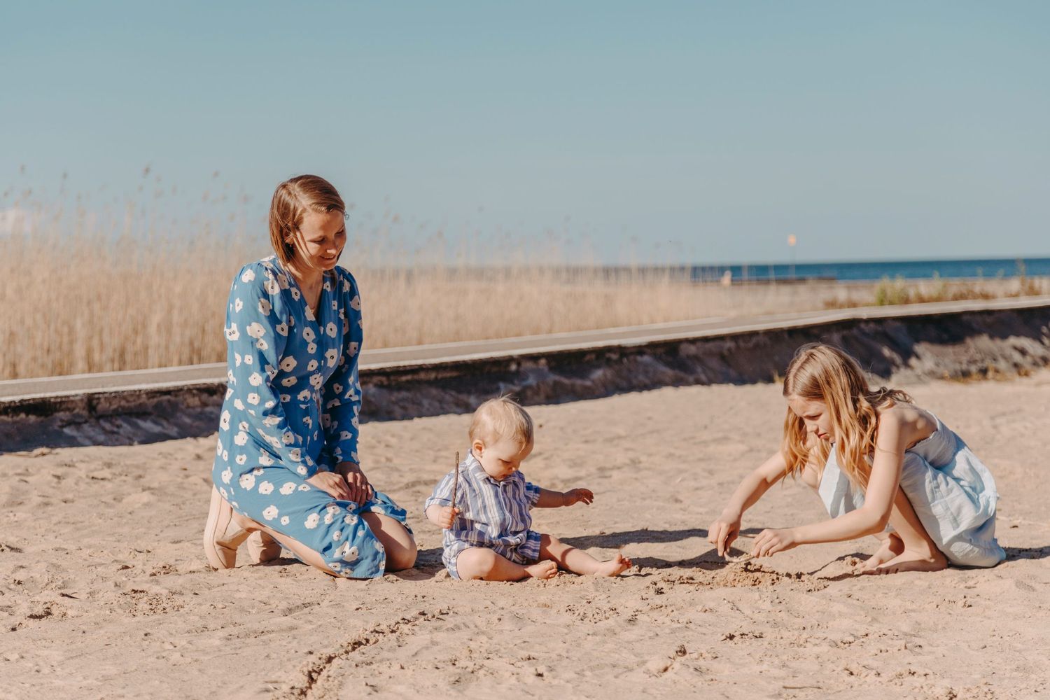 Three people sitting together on a sandy beach with tall grass and ocean in the background on a sunny day.
