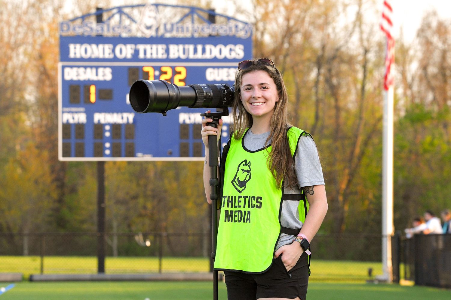 A sports photographer wearing a neon media vest stands by a scoreboard at a Bulldogs stadium.