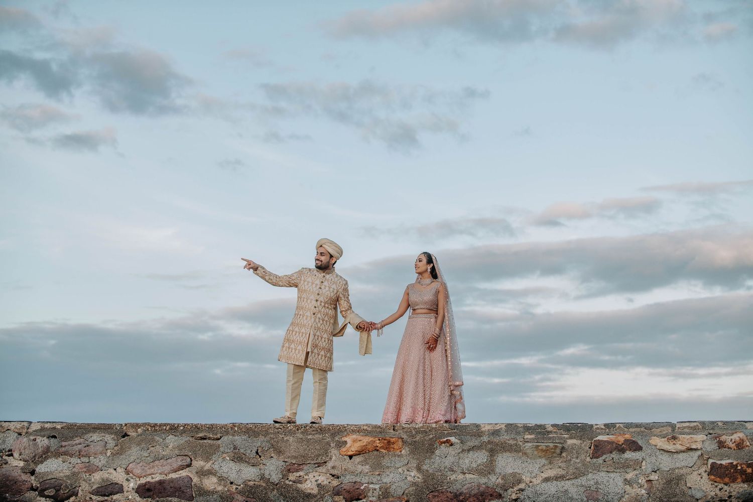 Couple in elegant attire stands on stone wall against cloudy sky at sunset holding hands and gesturing.
