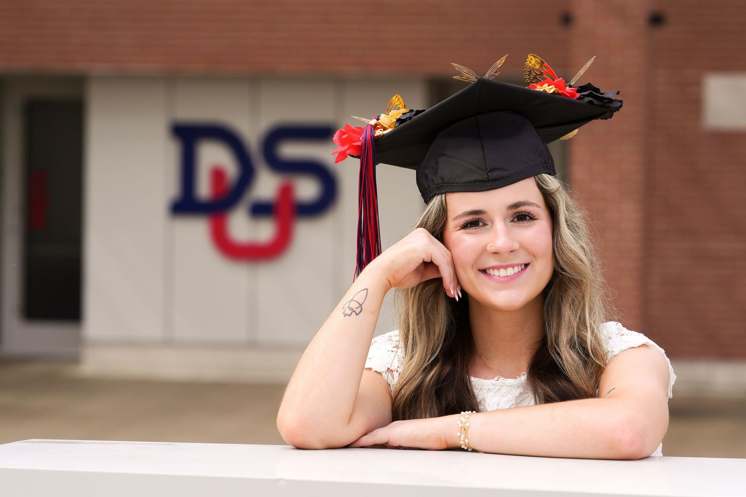 A graduate in cap and gown poses casually by a DeSales University sign.