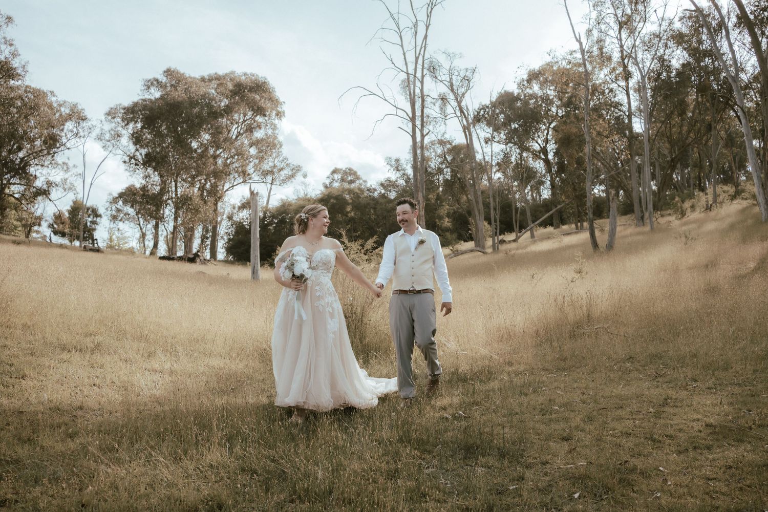 A couple walks hand in hand through an autumn field with eucalyptus trees in the background on their wedding day.