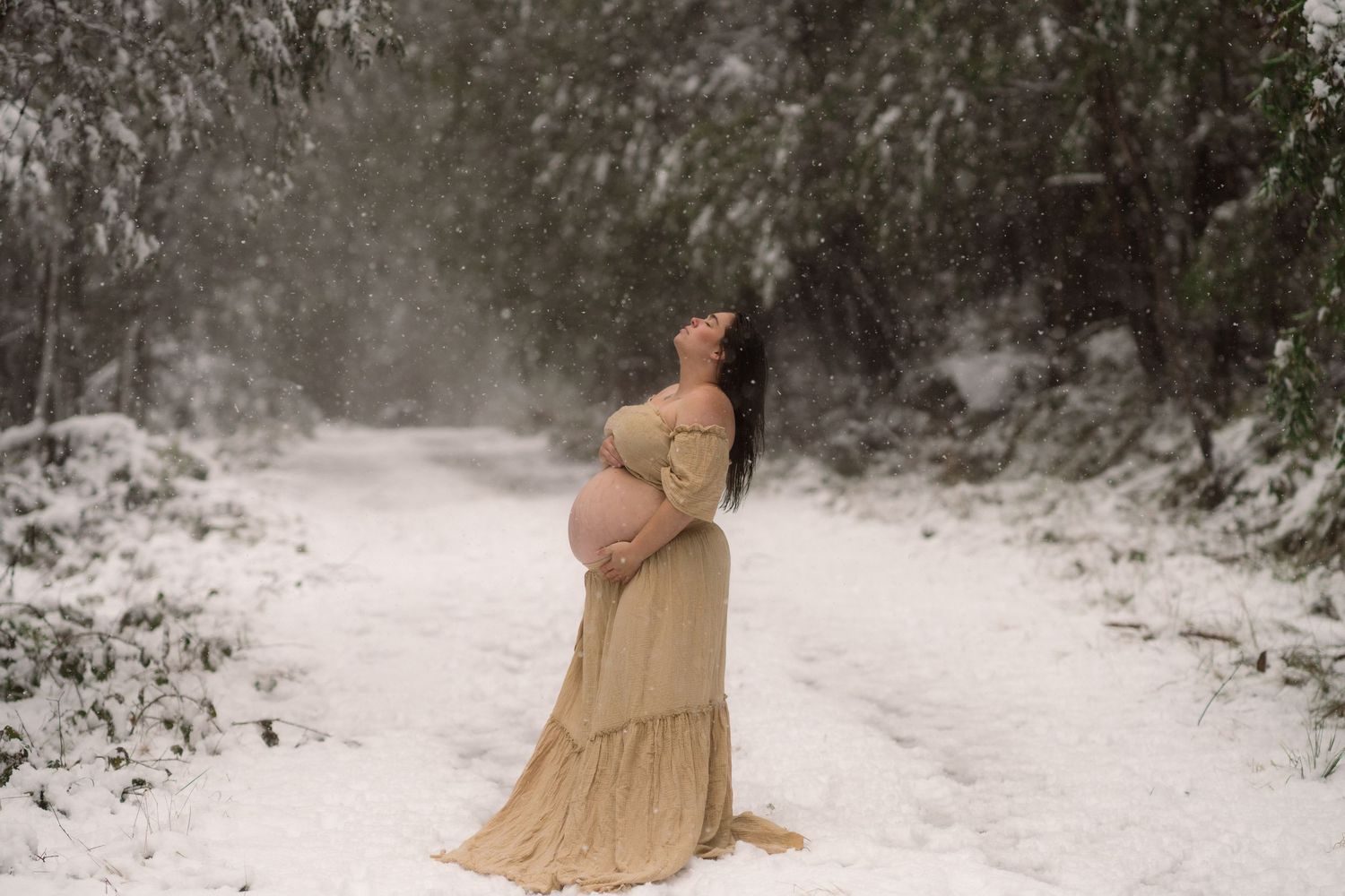 A pregnant woman in a flowing beige dress stands on a snowy path surrounded by frost-covered trees.