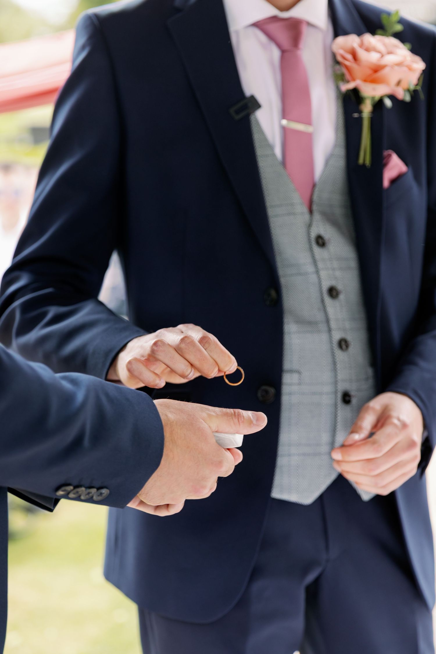 A navy-suited person holds and shows off their ring and pink boutonniere at a wedding.
