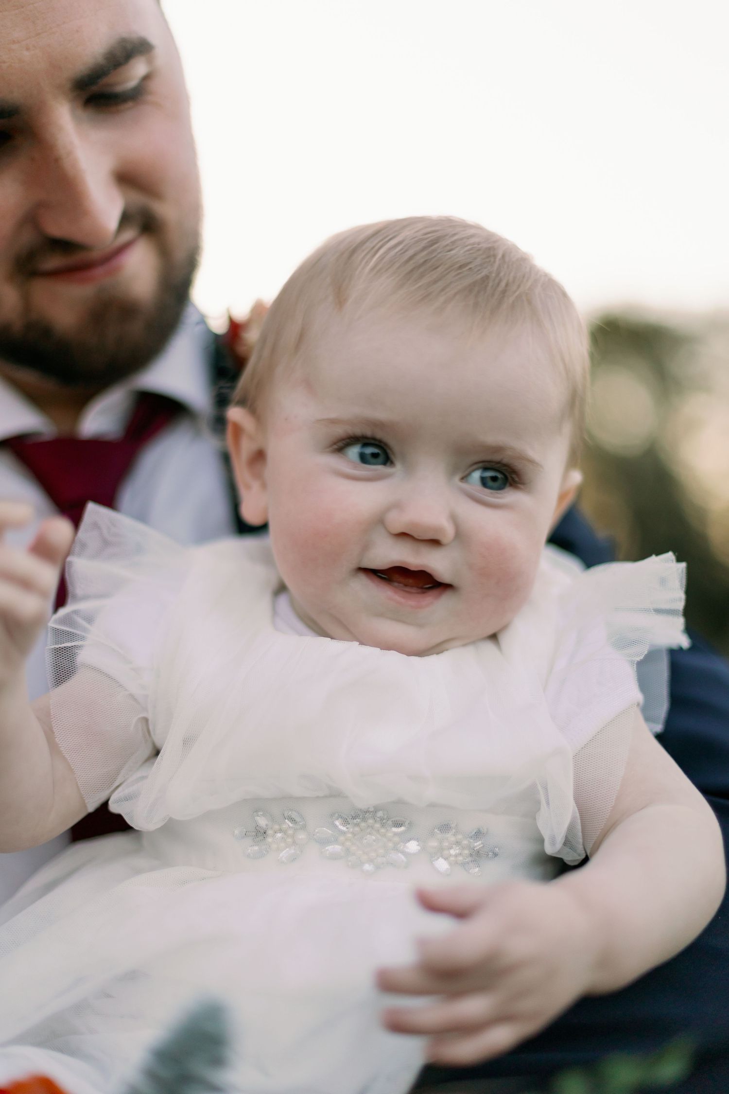 A baby in a light pink dress with ruffles and a sparkly sash smiles while being held by someone in formalwear.