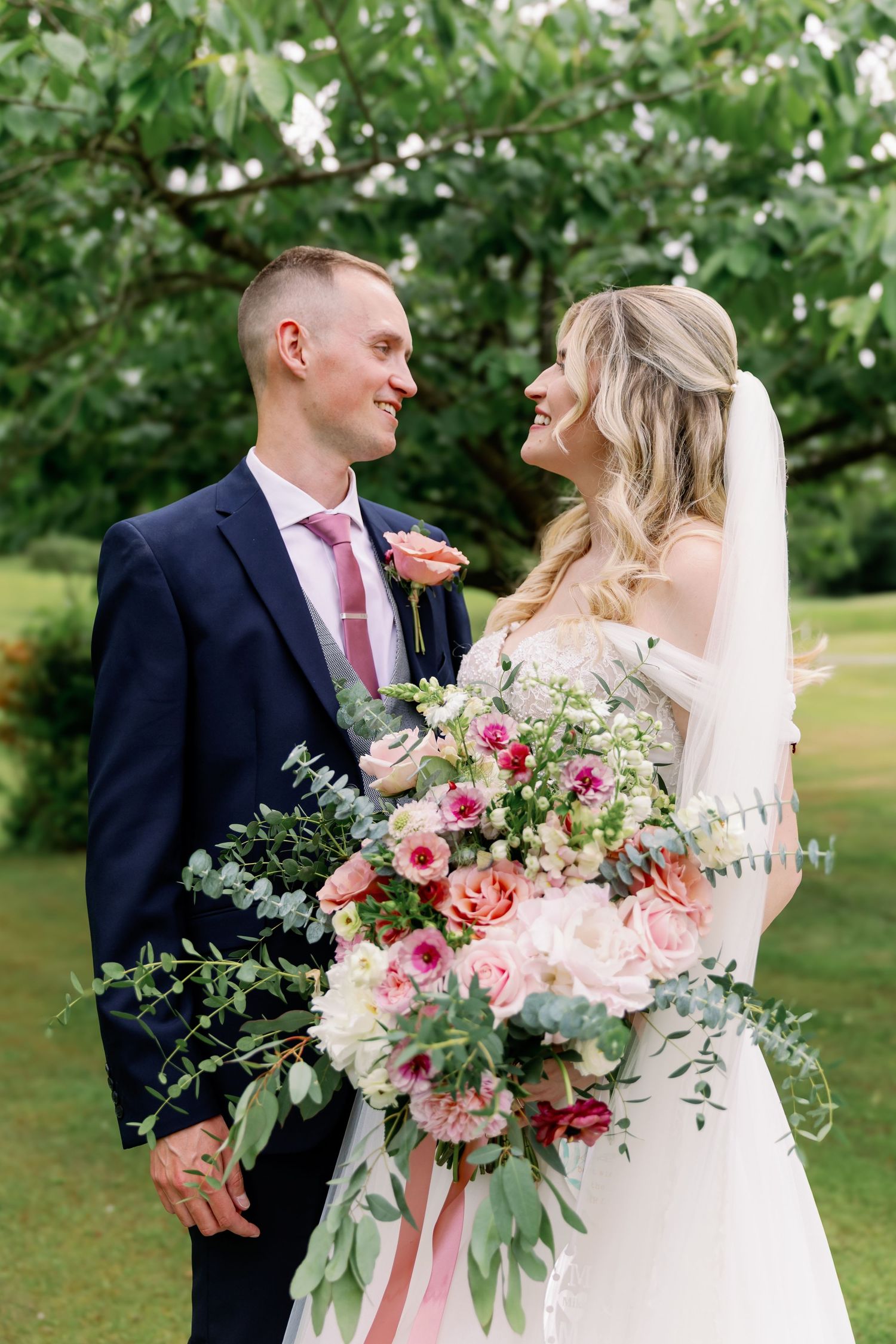 A happy couple on their wedding day share a loving moment surrounded by greenery with a stunning pink floral bouquet.