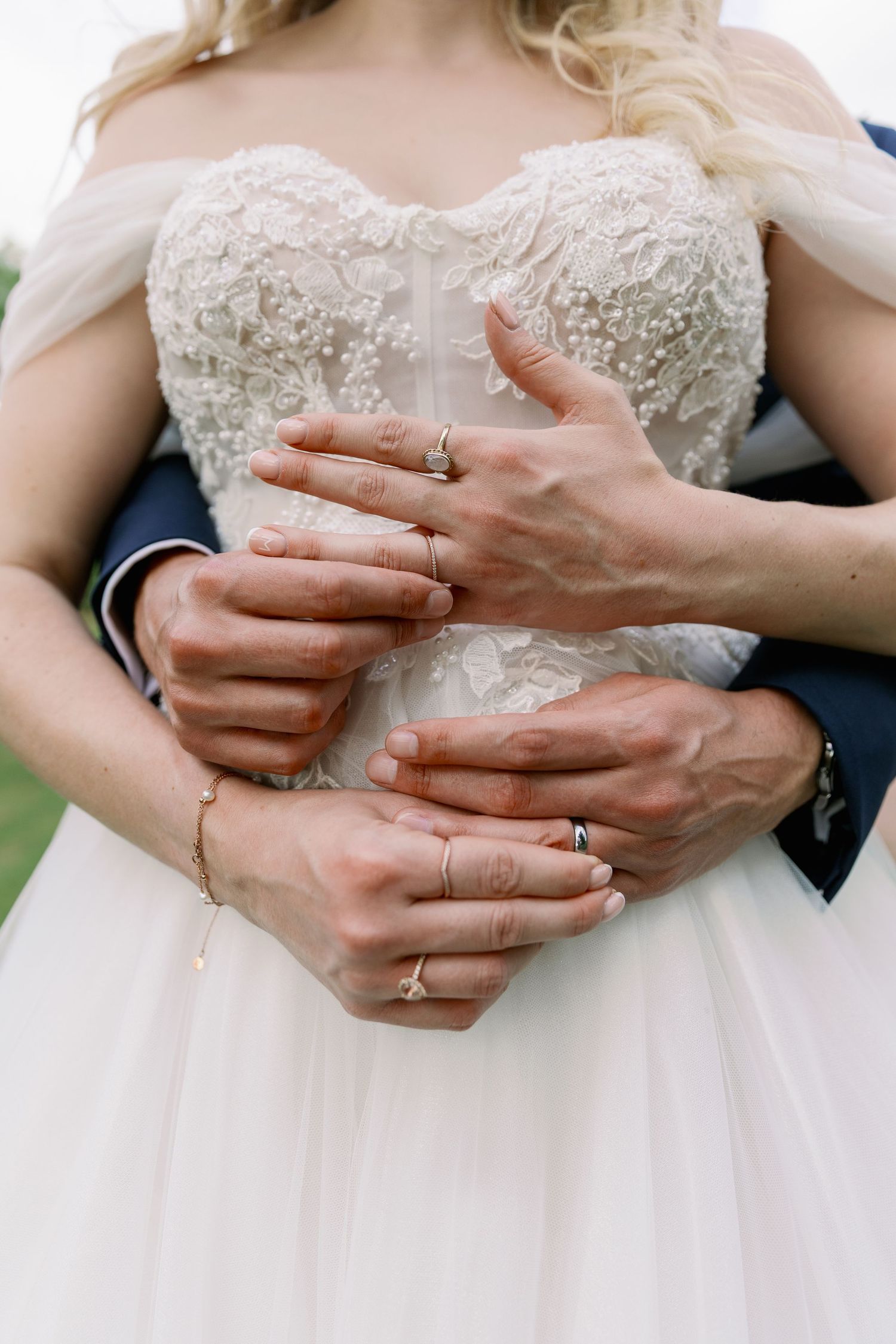 Close-up of hands wearing wedding rings embracing around delicate lace wedding dress bodice.
