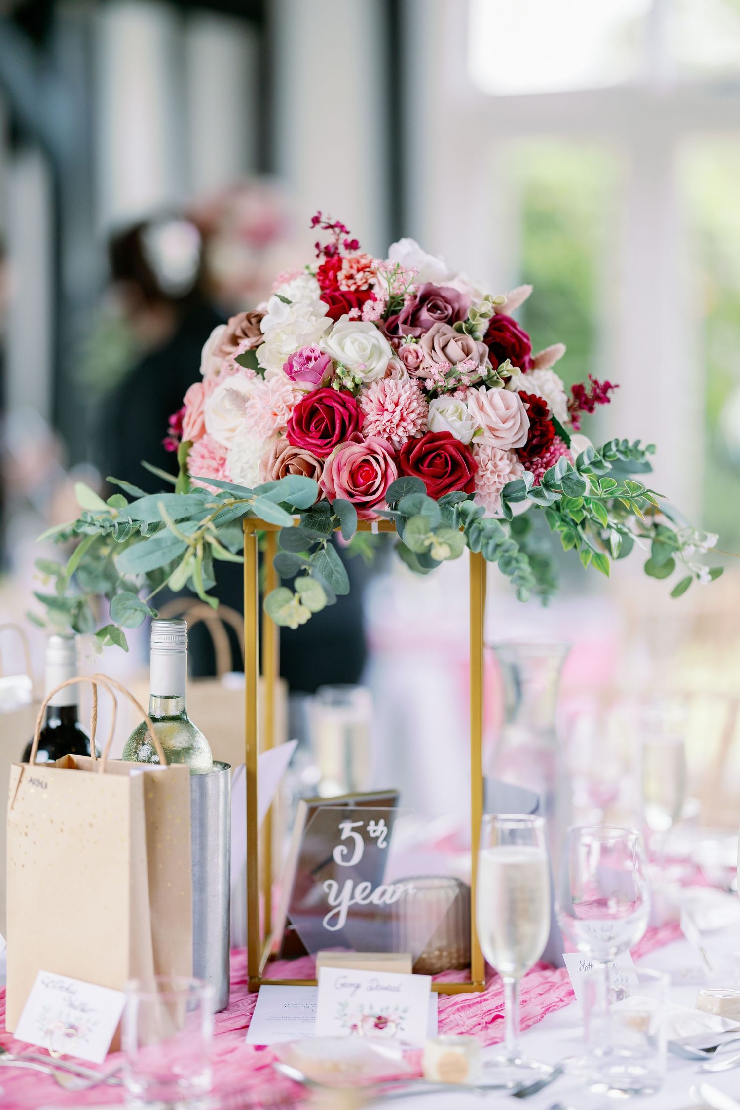 Elegant wedding table centerpiece with pink and red roses on gold stand next to wine bottles and place settings.