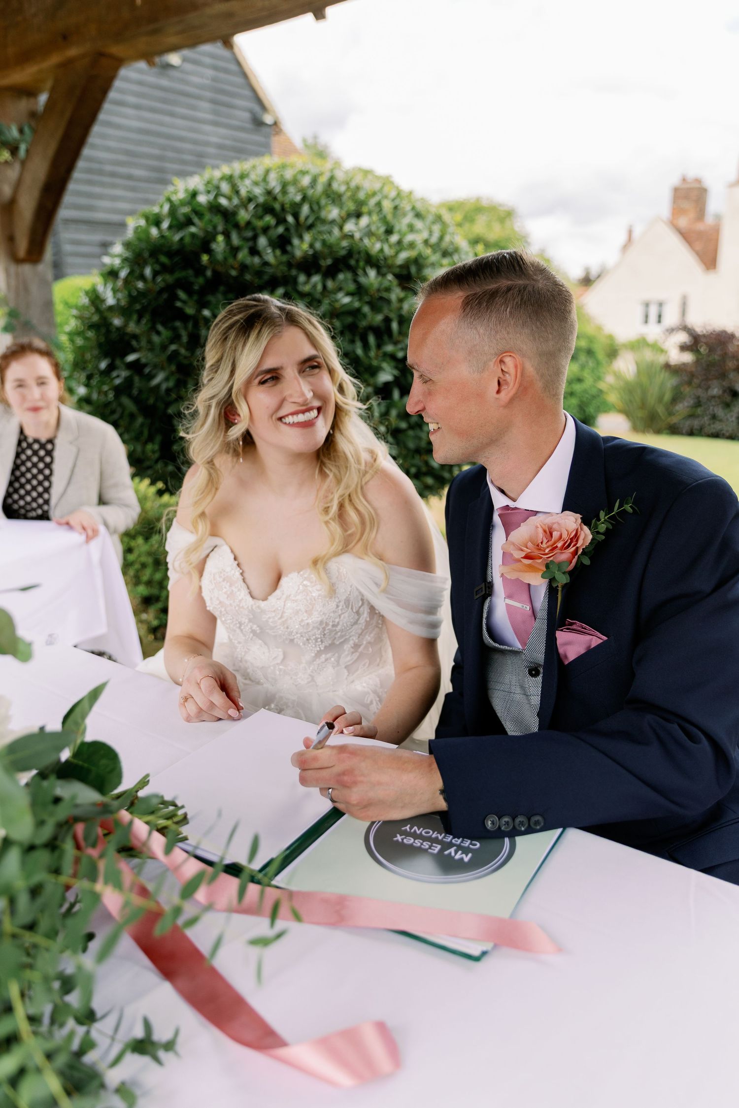 Couple signing marriage documents at outdoor wedding ceremony in sunny garden setting.