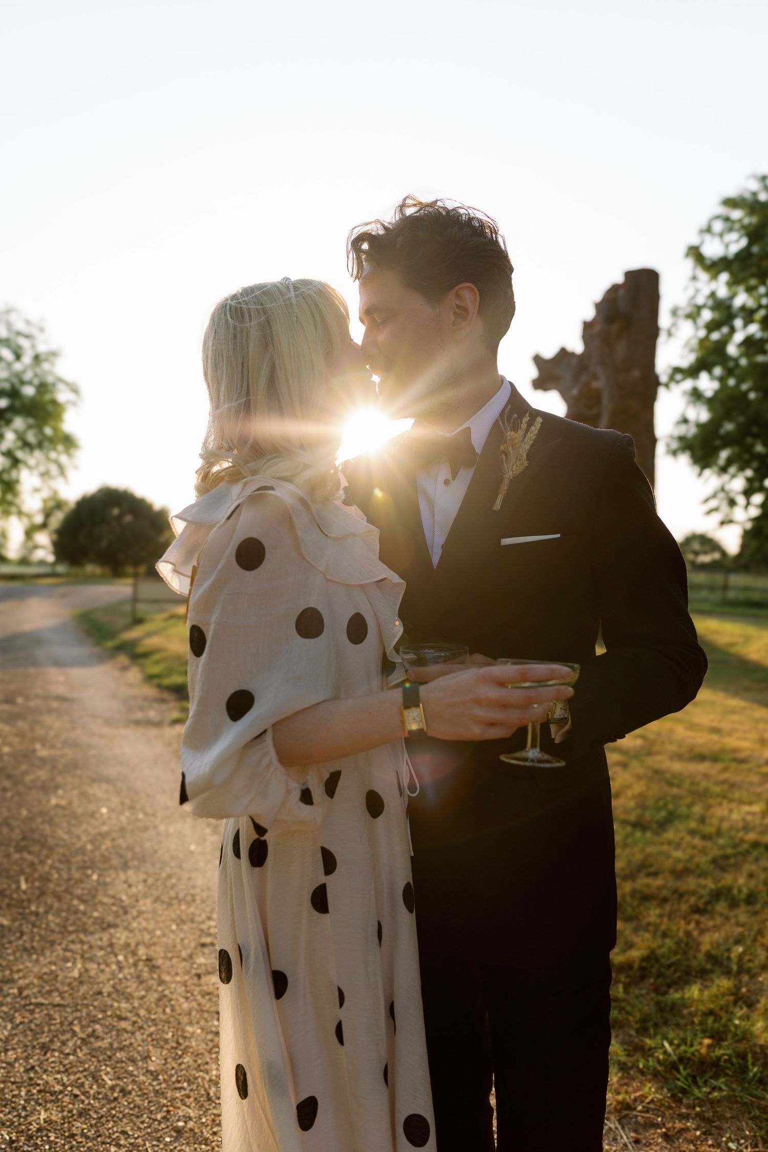 Couple in formal attire embrace on a path at sunset, with the woman wearing a polka dot dress and man in dark suit.