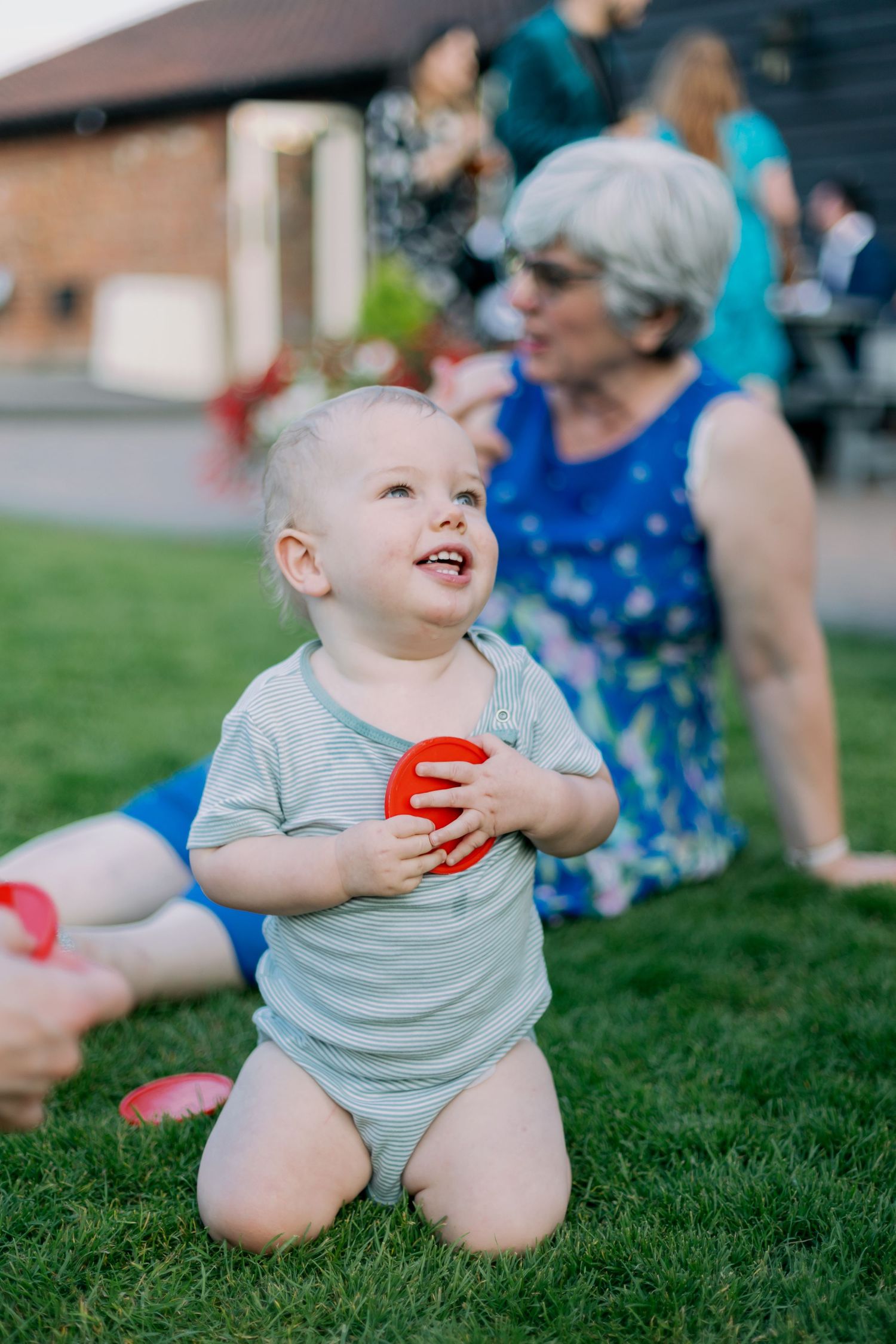 Young child in gray onesie plays with red toy while sitting on grass during outdoor gathering.