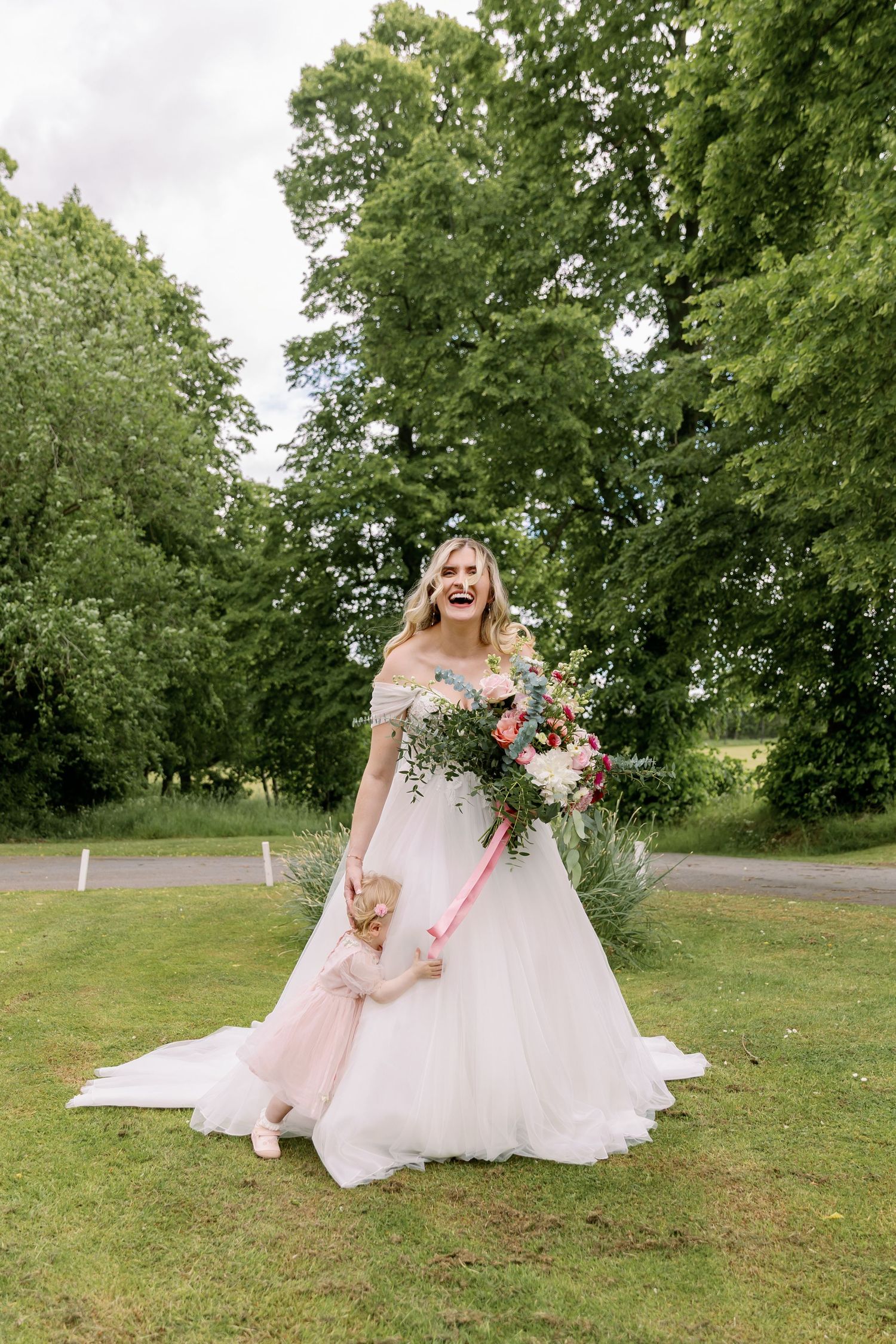 A bride in a flowing white off-shoulder wedding gown holds a cascading bouquet while standing on lush green grass.